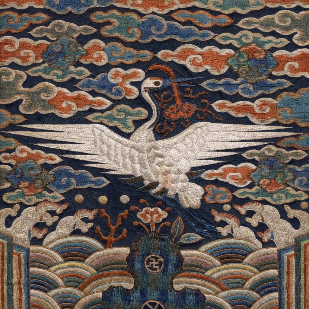 Una gru bianca con le ali spiegate è ricamata tra nuvole e onde colorate su uno sfondo tessile scuro, evocando l'eleganza e la tradizione che scoprirete durante un viaggio Corea o un Gran Tour della Corea.