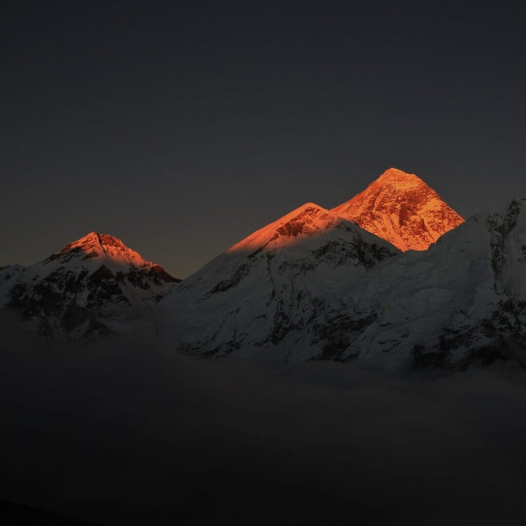 Le cime innevate delle montagne, illuminate dai raggi arancioni dei raggi del sole, si ergono sopra le nuvole nella Valle degli Dèi, contro un cielo scuro: un momento indimenticabile di ogni avventura del Tour Nepal.