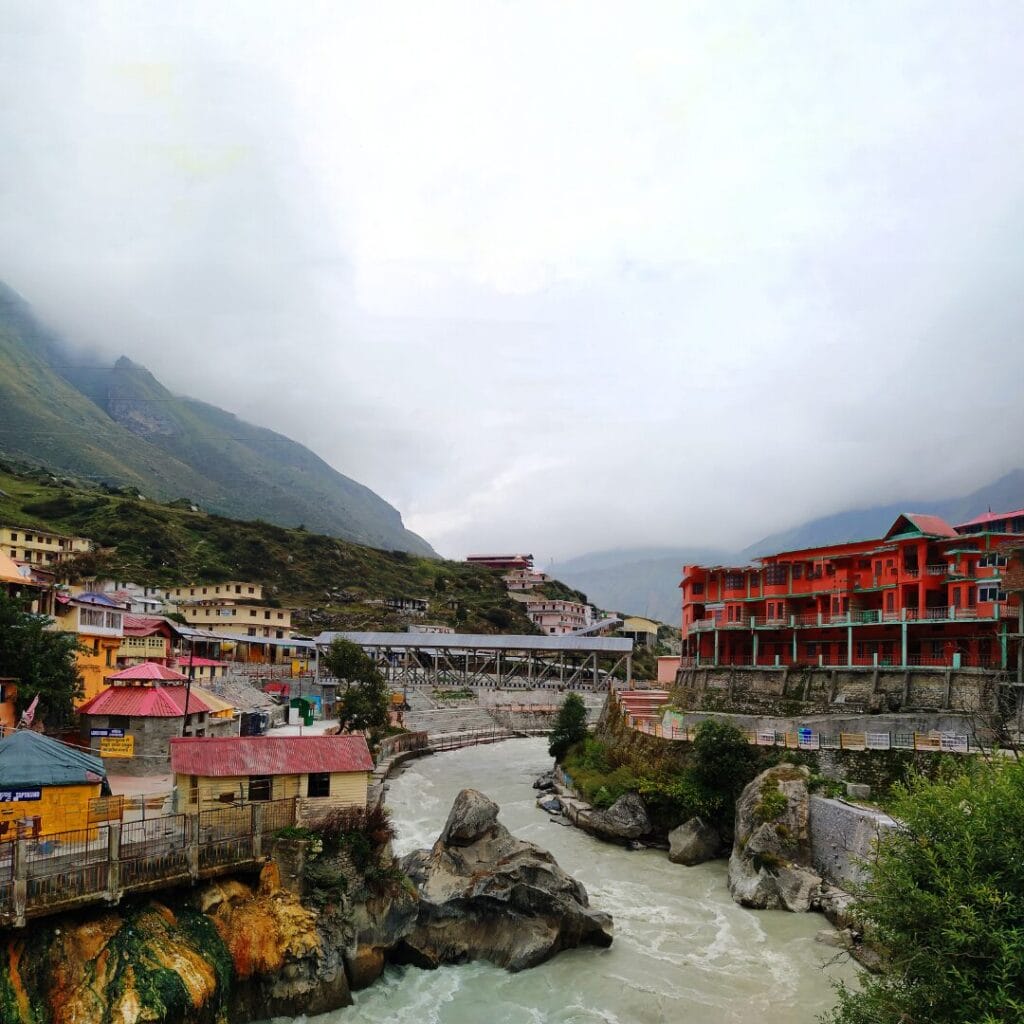 Un fiume scorre attraverso una cittadina di montagna in Nepal con edifici colorati e un ponte di metallo, circondata da colline nebbiose e cieli nuvolosi, catturando il fascino sereno spesso visibile durante il Festival di Dashain.