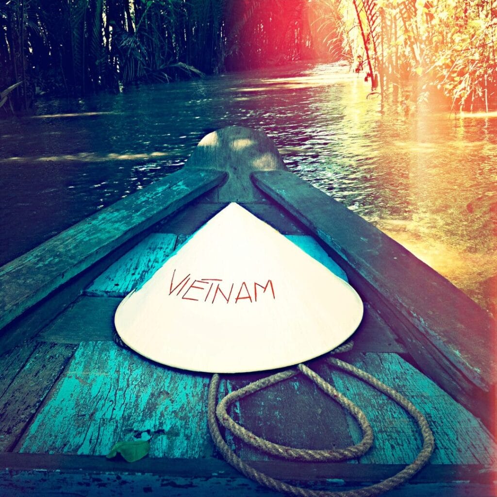 Un cappello conico con la scritta "Vietnam" è appoggiato su una barca di legno durante un Gran Tour del Vietnam, alla deriva lungo uno stretto fiume soleggiato e circondato da una vegetazione lussureggiante.