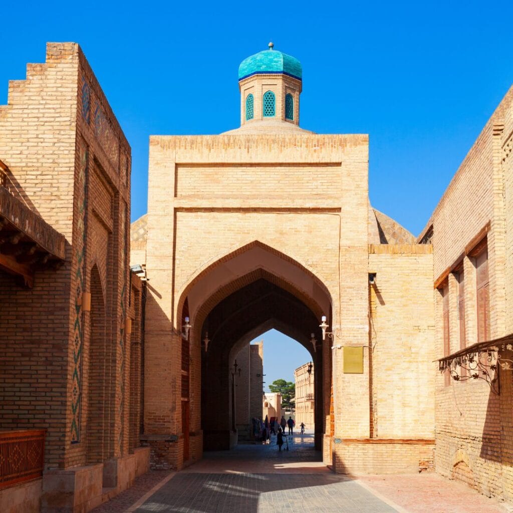 Uno storico arco in mattoni con una cupola blu in cima, fiancheggiato da edifici, sotto un cielo azzurro e limpido, con persone che camminano lungo il passaggio: uno spettacolo iconico di qualsiasi tour in Uzbekistan.