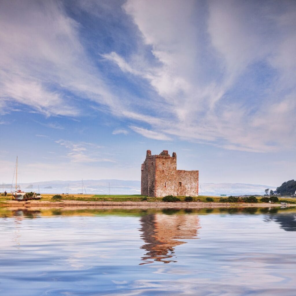 Un castello di pietra si erge su una riva erbosa nelle Highlands, riflesso nelle acque calme sotto un cielo parzialmente nuvoloso.