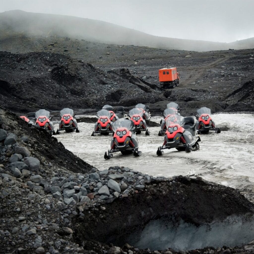 Un gruppo di motoslitte rosse è parcheggiato sul terreno roccioso e vulcanico di Sud, Isole Vestman in Islanda, vicino a un fiume, con un veicolo arancione sullo sfondo sotto un cielo nuvoloso.
