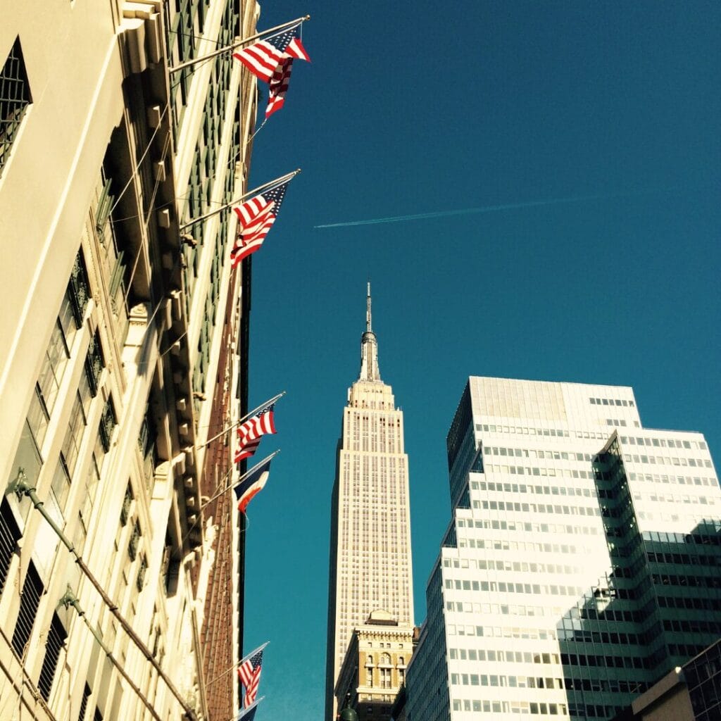 Vista dell'Empire State Building e dei grattacieli circostanti di New York City, con numerose bandiere americane su un edificio in primo piano: uno scenario mozzafiato da ammirare durante un tour di New York con un accompagnatore dall'Italia.