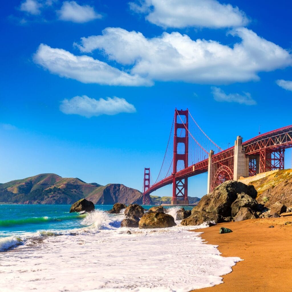 Il Golden Gate Bridge si estende sull'acqua azzurra ed è visibile da una spiaggia sabbiosa, con rocce in primo piano e colline sullo sfondo, sotto un cielo azzurro brillante: una tappa iconica lungo il Tour del Golden West.
