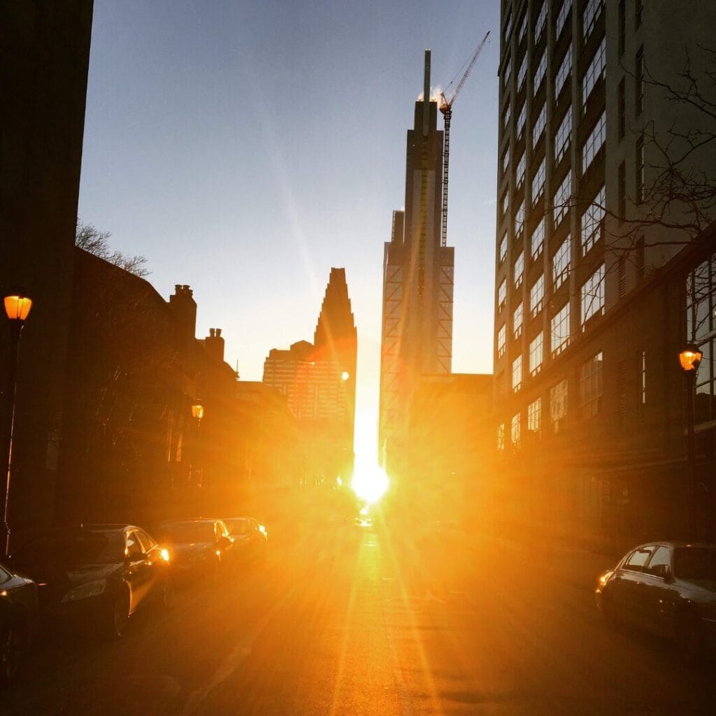 Una strada cittadina al tramonto con alti edifici su entrambi i lati, auto parcheggiate lungo la strada e la luce del sole che splende direttamente verso la telecamera: come una scena di un'avventura al Tour Splendore dell'Est o una vivace serata sulle cascate del Niagara.