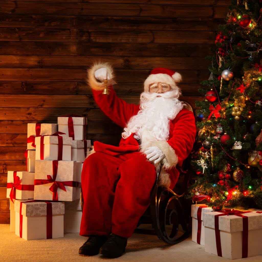 Babbo Natale è seduto su una sedia accanto a un albero di Natale decorato con l'Incanto Boreale, con in mano un campanello, circondato da regali incartati con nastri rossi e su uno sfondo di legno.