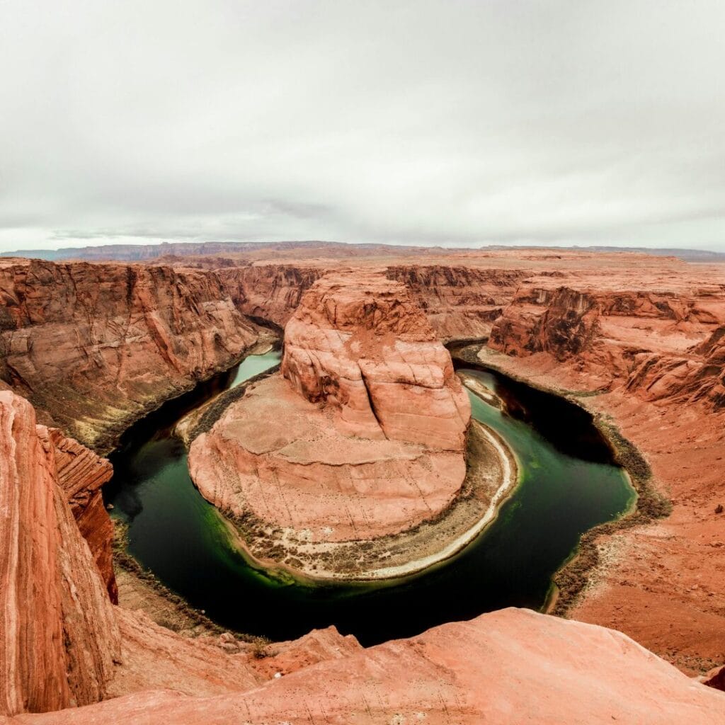 Un'ampia vista di Horseshoe Bend, dove il fiume Colorado si curva attorno a una formazione rocciosa di arenaria sotto un cielo nuvoloso in un paesaggio desertico, fondendo la bellezza naturale con la modernità e le tradizioni delle iconiche capitali del turismo.