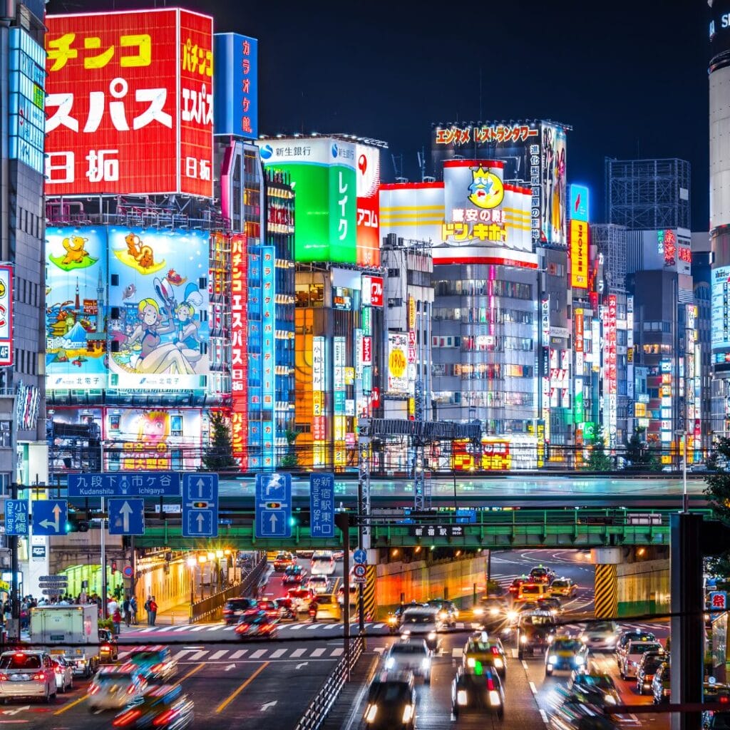Strada trafficata della città di notte in Giappone, con traffico intenso e numerosi cartelloni pubblicitari e insegne al neon illuminati sugli edifici alti, che catturano la vibrante energia della vita del centro urbano, uno scenario da esplorare durante il Foliage 2025.