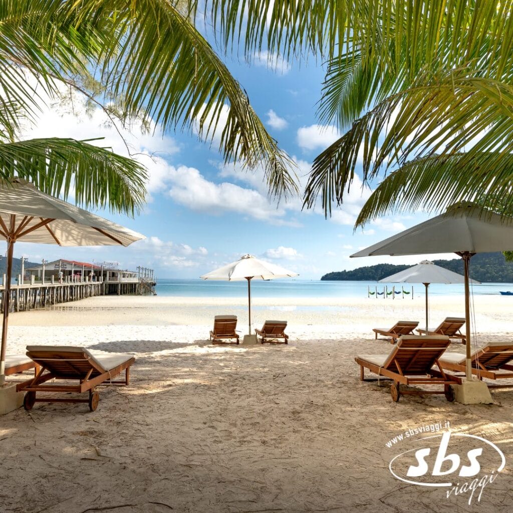 Lettini e ombrelloni in legno su una spiaggia tropicale di sabbia a Phuket, con palme e un molo sullo sfondo: perfetti per la vostra Estensione Mare durante un Tour Thailandia.