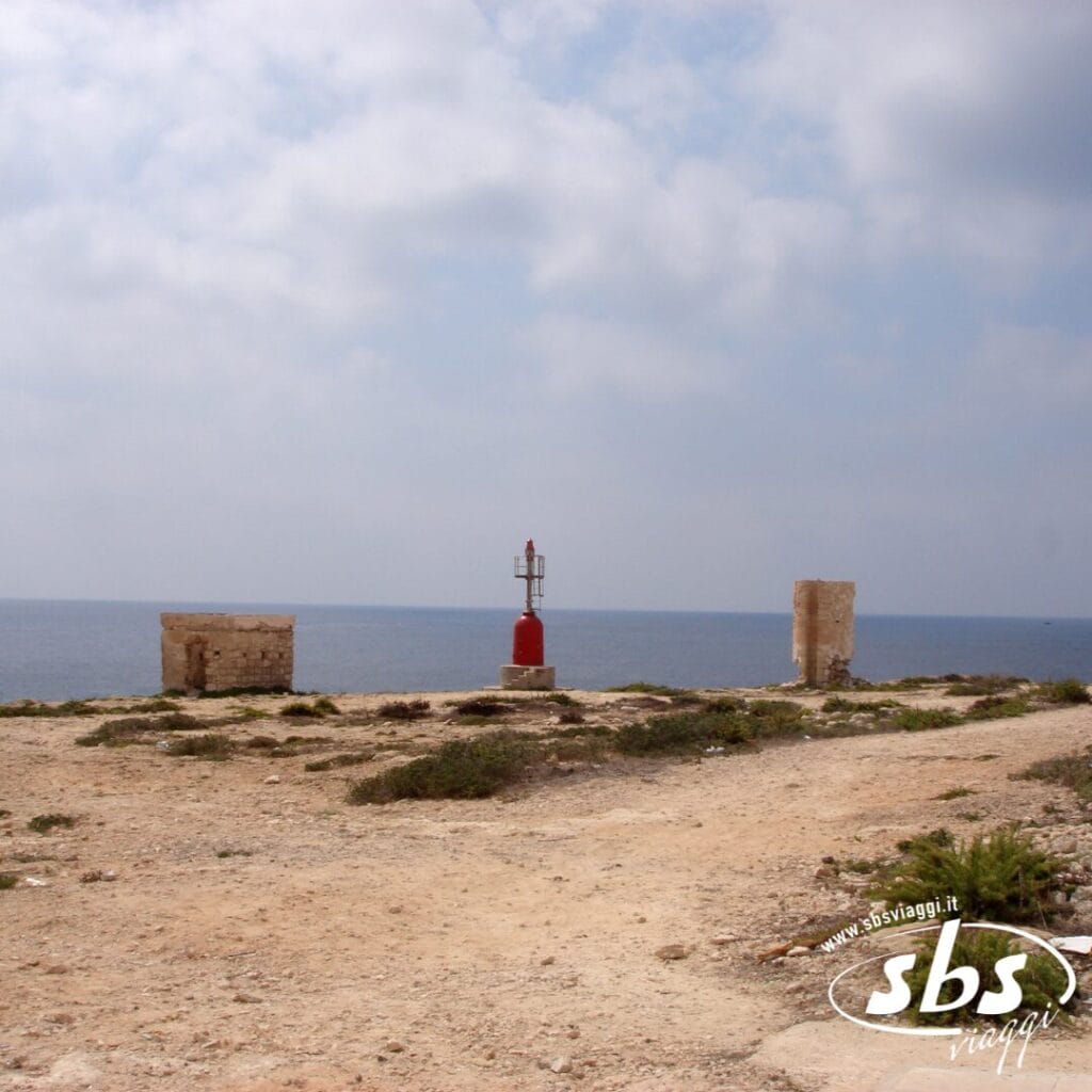 Un piccolo faro rosso si erge su una costa rocciosa vicino ad antiche rovine, con il mare e un cielo nuvoloso sullo sfondo: uno scenario suggestivo, perfetto per i viaggiatori in cerca di esperienze uniche in hotel o villaggi in riva all'acqua.