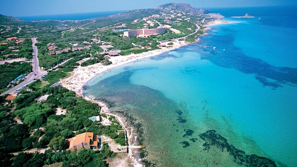 Vista aerea di una costa con acque turchesi, spiaggia sabbiosa, verdi colline con edifici e un tocco di tranquillità.
