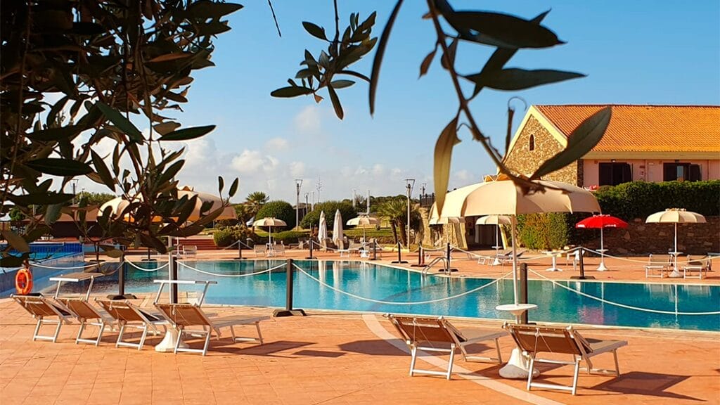 Piscina soleggiata con sedie a sdraio e ombrelloni, circondata dal verde e da un edificio con tetto in tegole: il luogo perfetto per rilassarsi in questo rifugio bozza automatica.