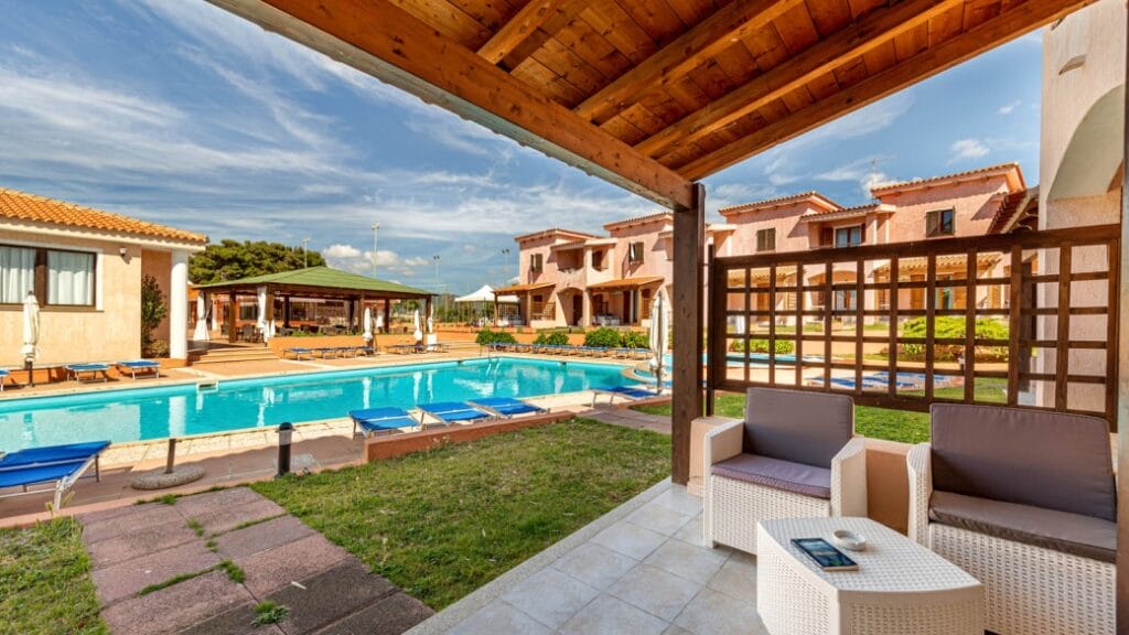 Patio coperto con sedie e vista sulla piscina, lettini prendisole ed edifici in stile mediterraneo sotto un cielo azzurro: perfetto per rilassarsi in questo ambiente da bozza automatica.