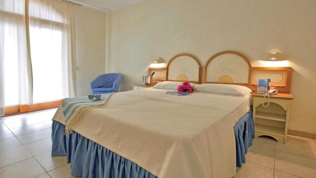 Luminosa camera d'albergo con letto matrimoniale, gonna blu, poltrona, comodini e fiori sul letto: uno spazio invitante che mette in risalto gli elementi di design Bozza automatica per un tocco moderno.