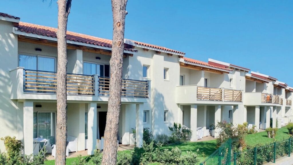 Un condominio bianco a due piani con balconi, circondato dal verde e dagli alberi sotto un cielo azzurro e terso: una Bozza automatica perfetta per una vita urbana tranquilla.