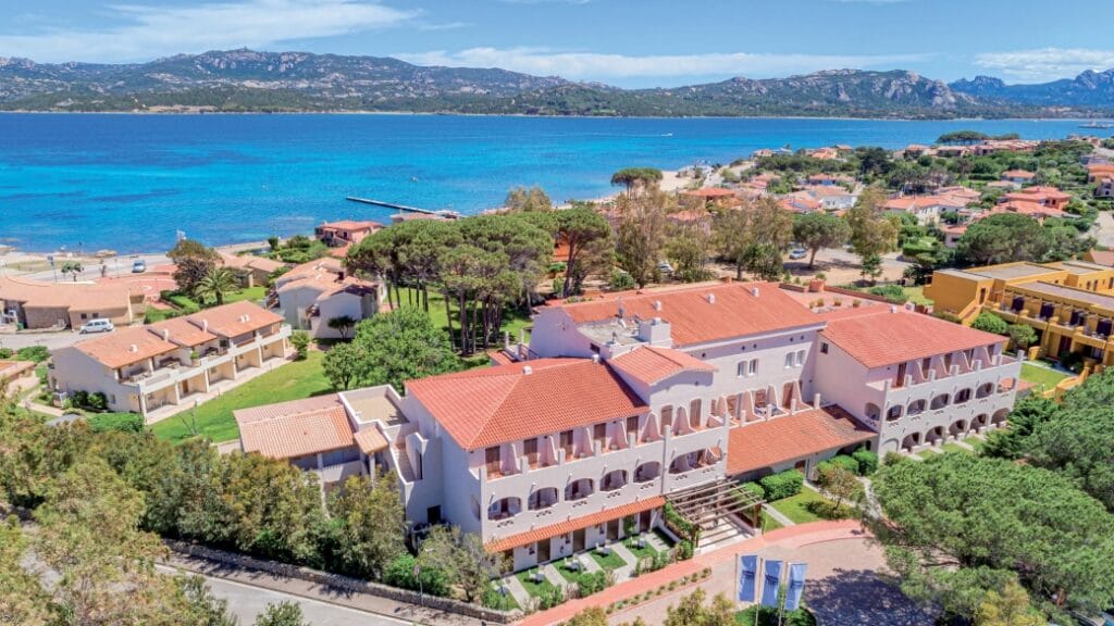 Un resort costiero con tetti di tegole rosse vicino al mare, circondato da alberi e vegetazione lussureggiante sotto un cielo azzurro: la perfetta fuga Bozza automatica.