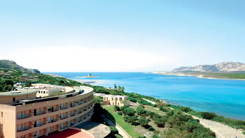 L'hotel Club Esse Roccaruja, dalla forma curva, si affaccia sul mare azzurro brillante nei pressi di Stintino, con verdi colline e una lontana costa rocciosa sotto un cielo limpido.
