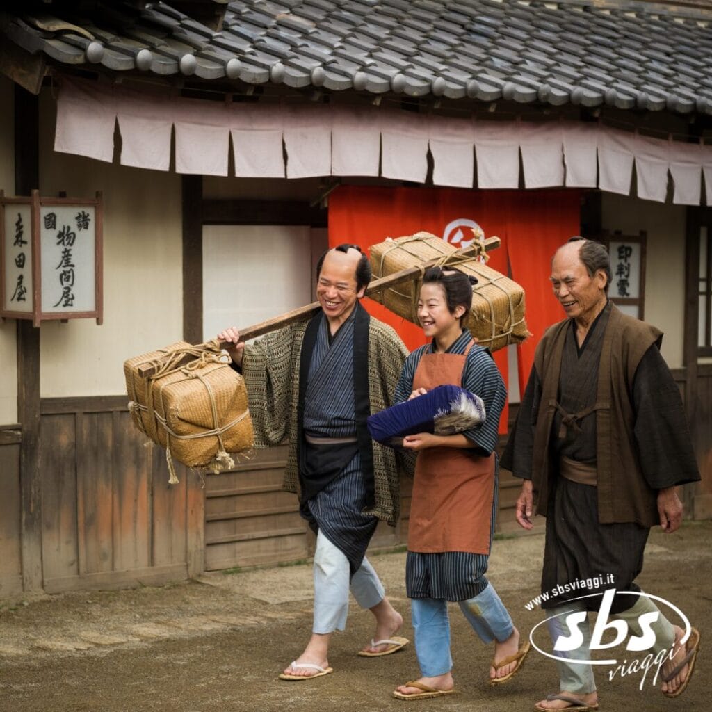 Tre persone in abiti tradizionali giapponesi camminano insieme fuori da un vecchio edificio in legno vicino a un onsen: due trasportano grandi fagotti e una tiene in mano un pacco incartato, catturando così il fascino senza tempo del Giappone.