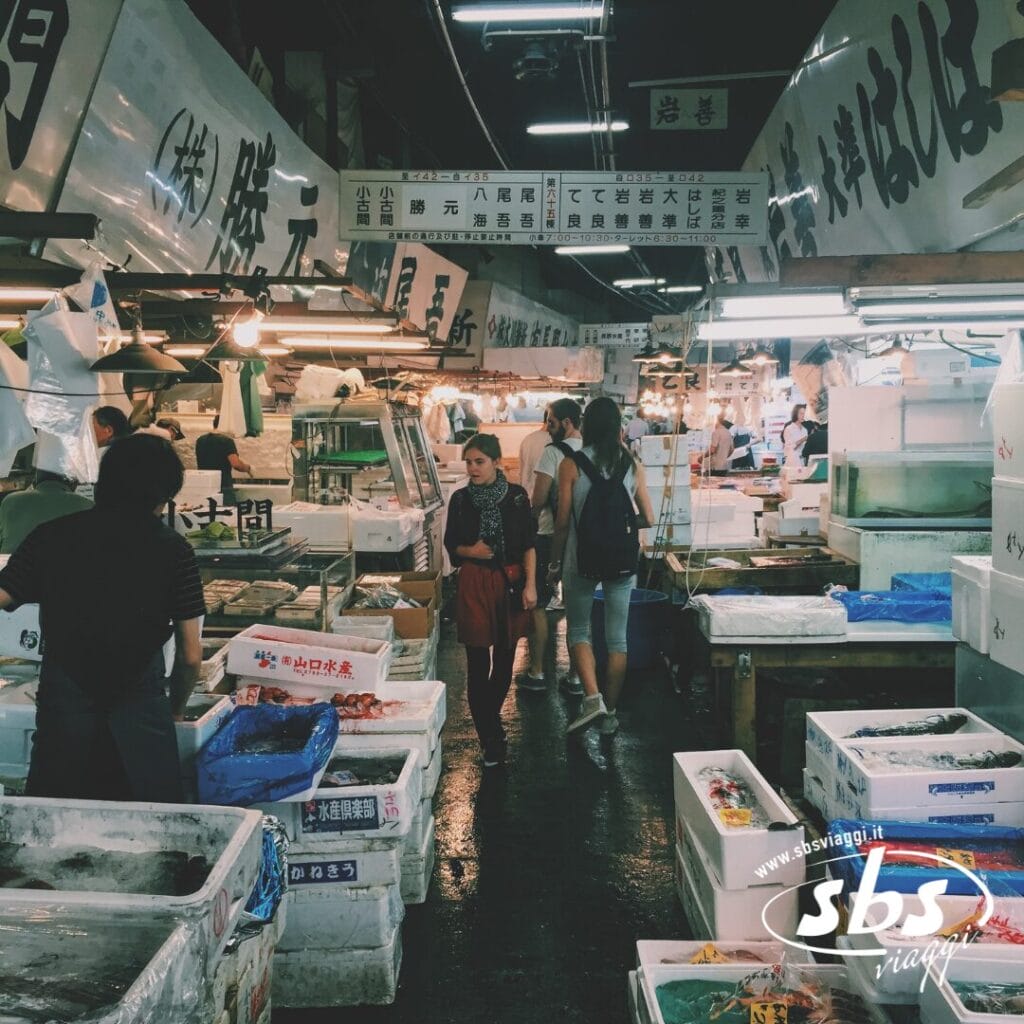La gente passeggia nel vivace mercato del pesce di Corea, con bancarelle che espongono frutti di mare in contenitori bianchi sotto luci intense e insegne giapponesi: una tappa vivace del Gran Tour della Corea.