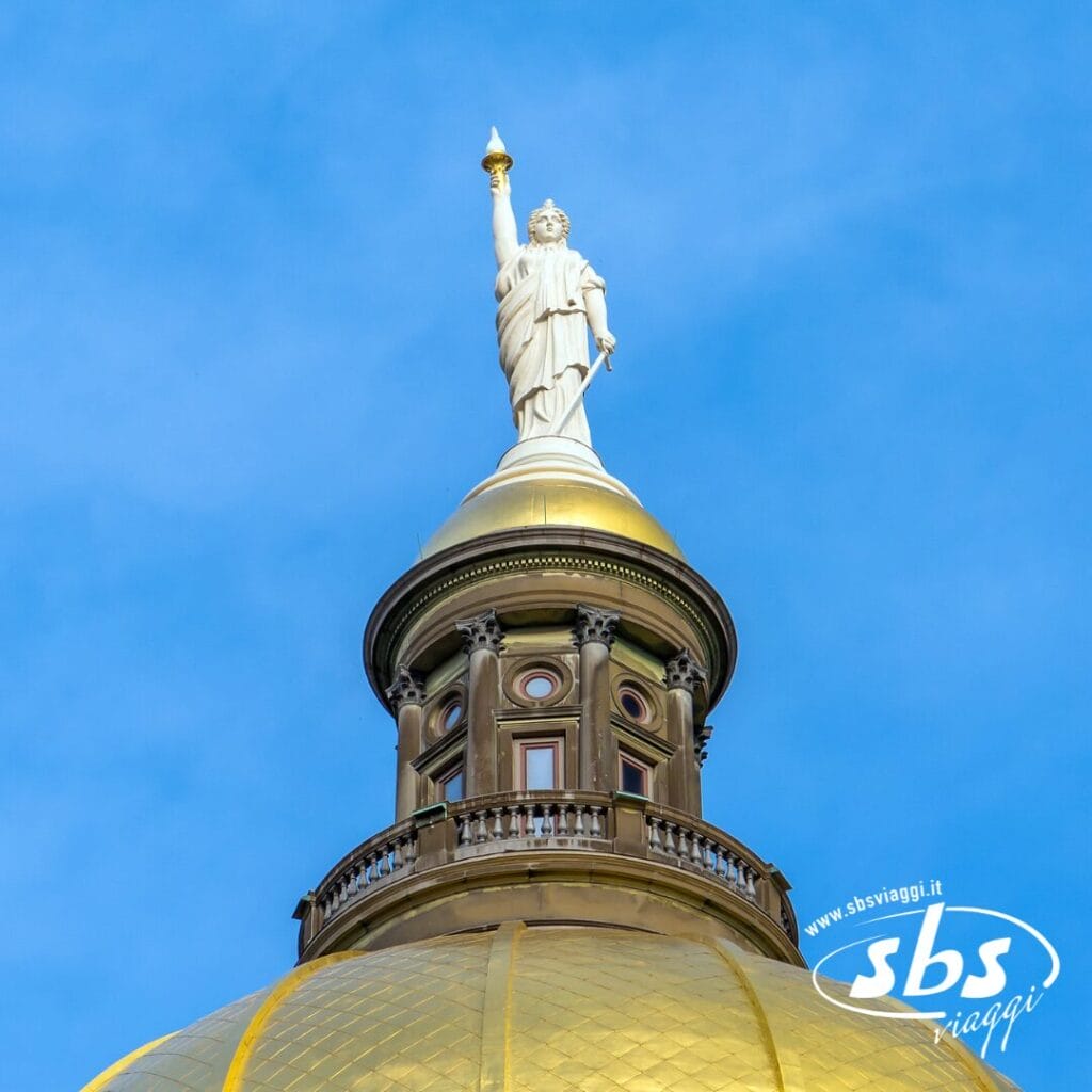 Primo piano di un edificio con cupola dorata in Georgia, sormontato da una statua bianca che regge una torcia, sullo sfondo di un cielo azzurro e terso.