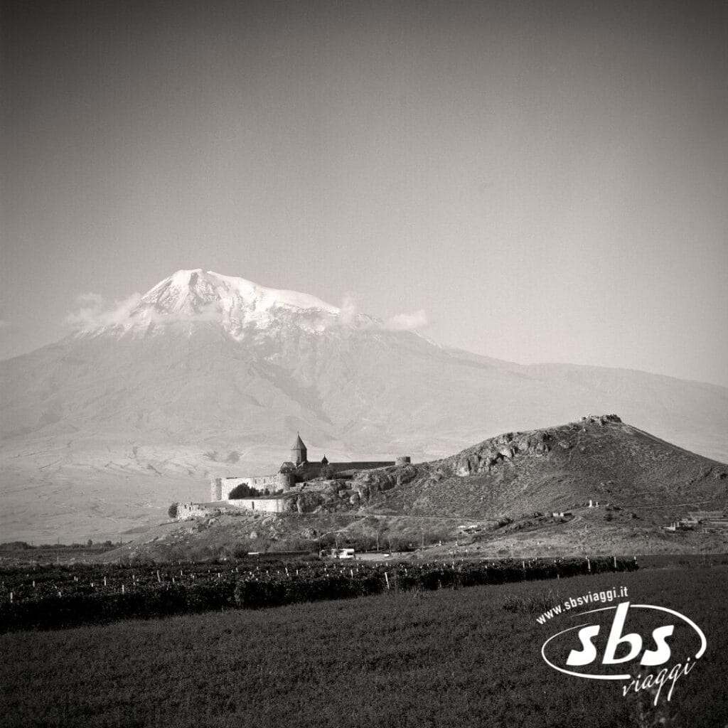 Una foto in bianco e nero mostra un monastero su una collina in Georgia, con una montagna innevata sullo sfondo. Il logo di
