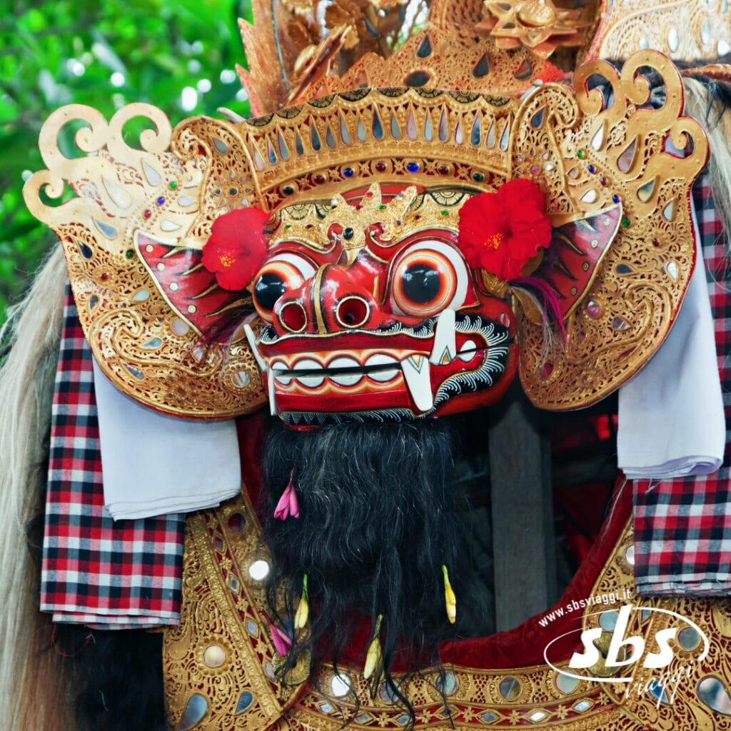 Una maschera tradizionale Barong con elaborati dettagli in oro, fiori rossi, tessuto a scacchi e denti sporgenti, fotografata all'aperto: uno sguardo ai segreti artistici di Bali per chi partecipa a un tour culturale.
