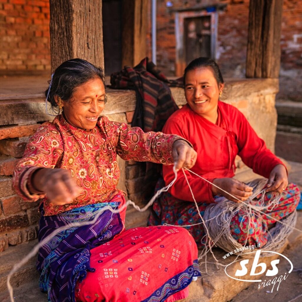 Due donne siedono su un portico rustico nella Valle degli Dei, sorridenti mentre lavorano con il filato. Una dipana i fili mentre l'altra osserva, godendosi un momento di pace all'aria aperta. Visitate il Nepal e scoprite scenari così sereni.
