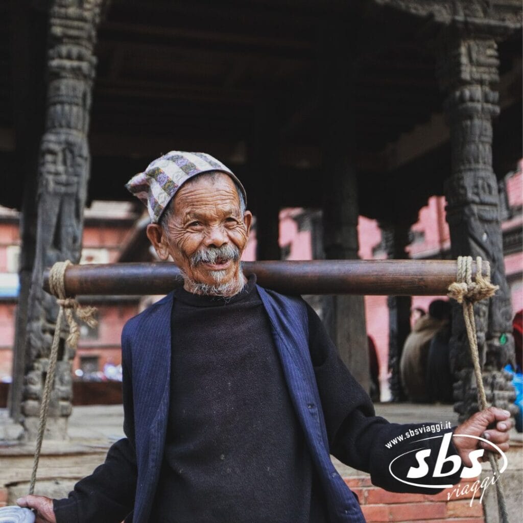 Un uomo anziano che indossa un cappello e un gilet decorati porta un palo di legno sulle spalle in un cortile nepalese con pilastri in pietra scolpita, evocando la ricca cultura ammirata durante un tour del Triangolo d'Oro.
