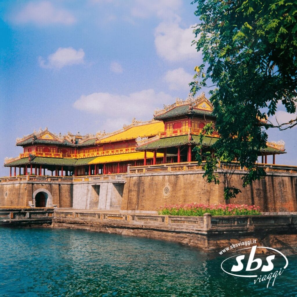 Un antico palazzo giallo e rosso con il tetto decorato si erge accanto a un fossato sotto un cielo azzurro, parzialmente incorniciato dai rami degli alberi: una tappa obbligata di qualsiasi tour in Vietnam.