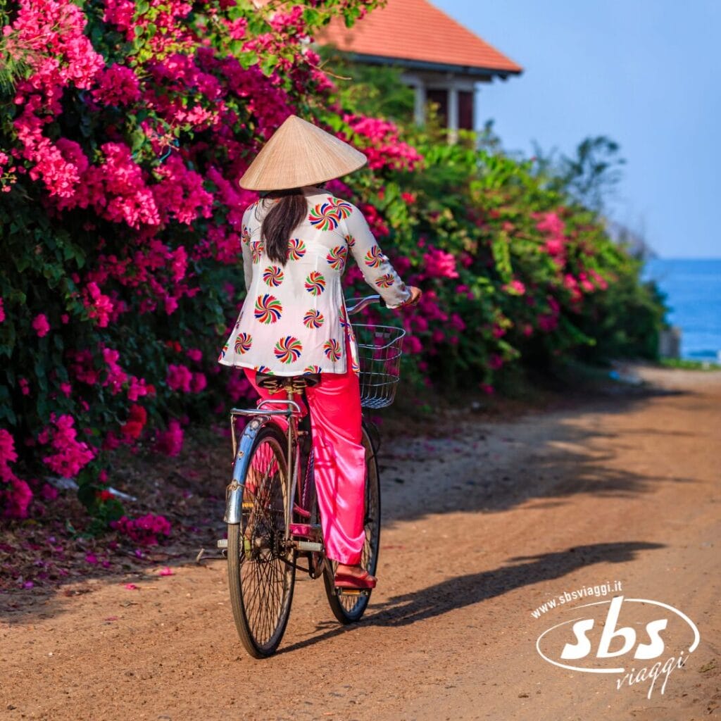 Una persona con un cappello conico e abiti colorati percorre in bicicletta un sentiero sterrato costeggiato da fiori rosa in fiore, vicino a un edificio e al mare: un'istantanea perfetta di un'autentica esperienza di viaggio in Vietnam.