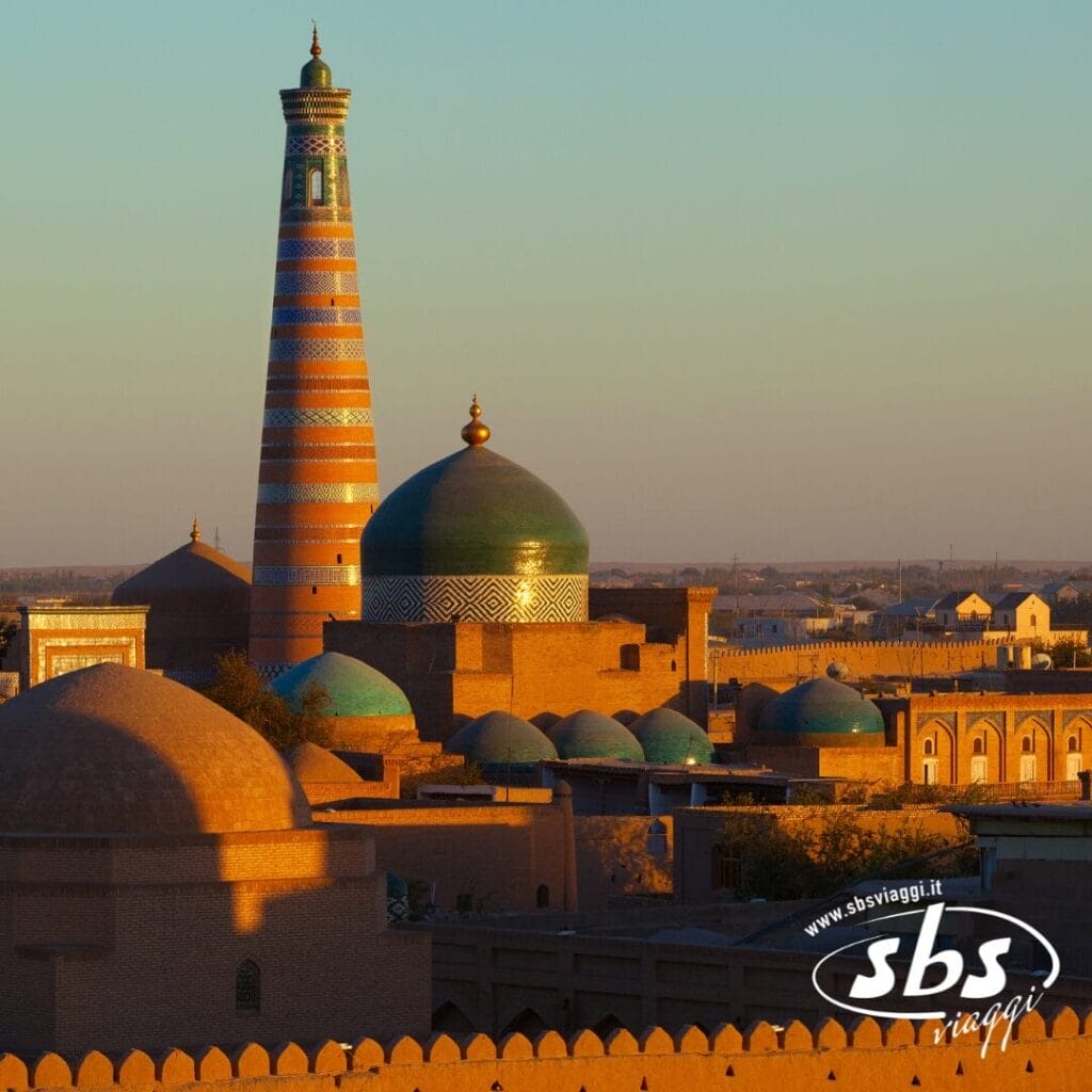 Vista soleggiata di edifici storici con cupole turchesi e un alto minareto a strisce a Khiva, in Uzbekistan, perfetta per il tuo prossimo tour in Uzbekistan con il logo SBS Viaggi nell'angolo.