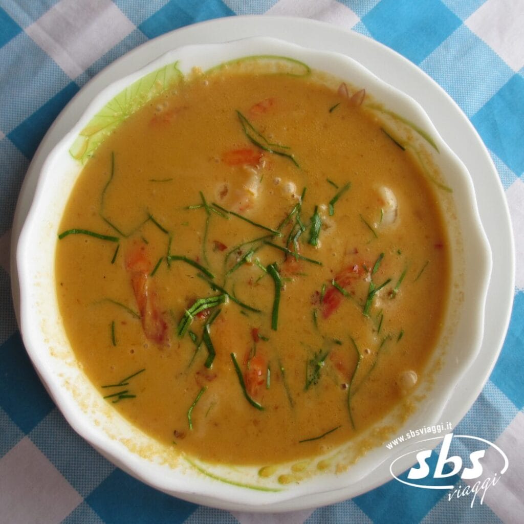 Una ciotola di cremosa zuppa all'arancia con erbe aromatiche e pezzi di pomodoro a vista, adagiata su un piatto bianco sopra una tovaglia a quadri bianchi e blu, evoca i sapori confortanti che spesso si possono scoprire durante un tour Grantour o Cambogia.