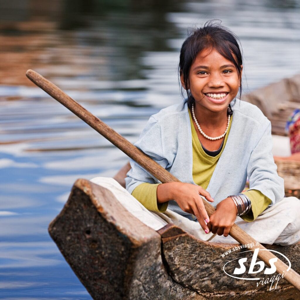 Una ragazza sorridente siede su una barca di legno sull'acqua, con una pagaia in mano. Indossa una giacca leggera e gioielli, circondata da cesti, catturando lo spirito autentico di un tour in Cambogia.