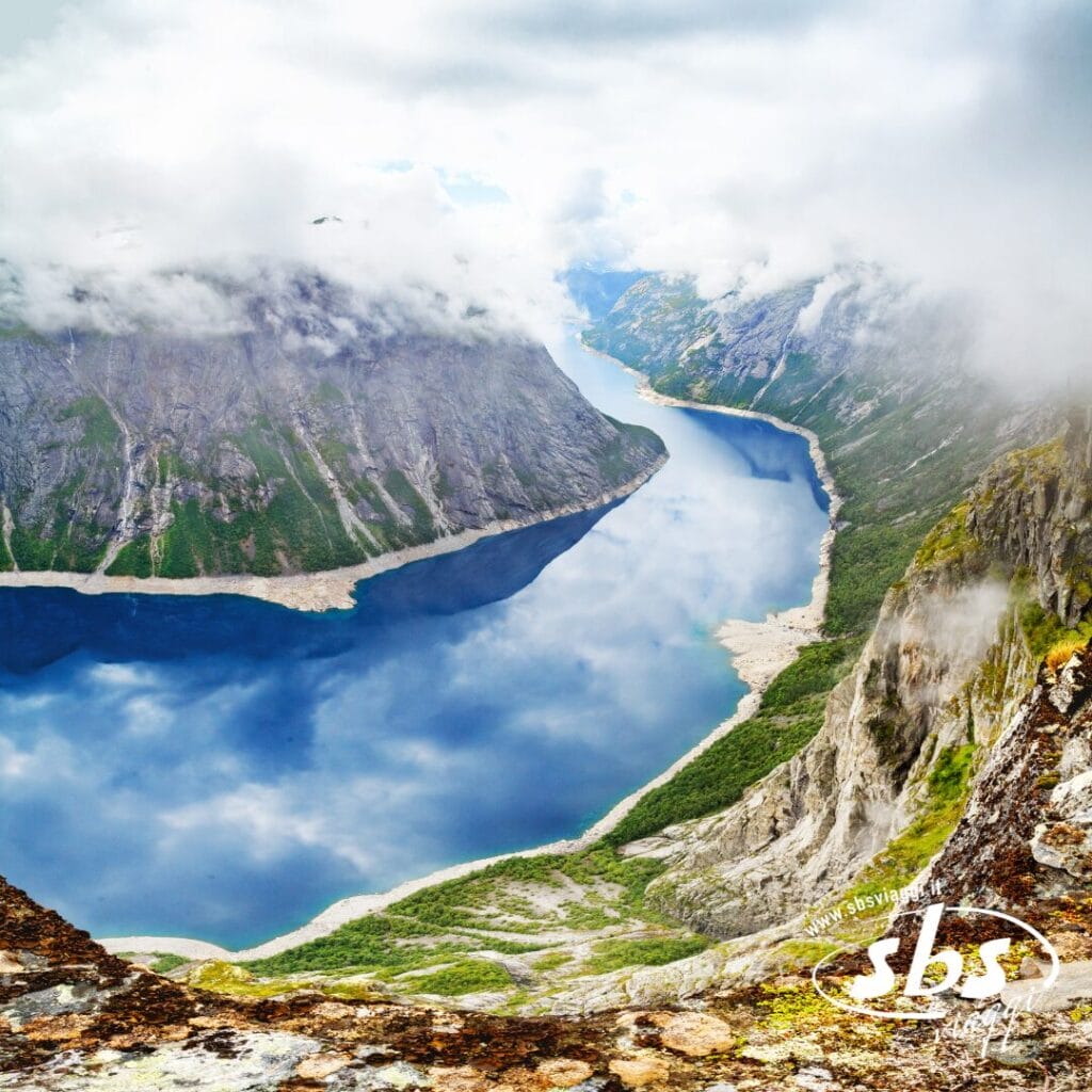 Un fiume azzurro e limpido serpeggia tra montagne ripide e rocciose ricoperte di verde, con nuvole che ne nascondono parzialmente le cime: uno scenario perfetto per un Tour Fiordi Meravigliosi con accompagnatore dall’Italia.