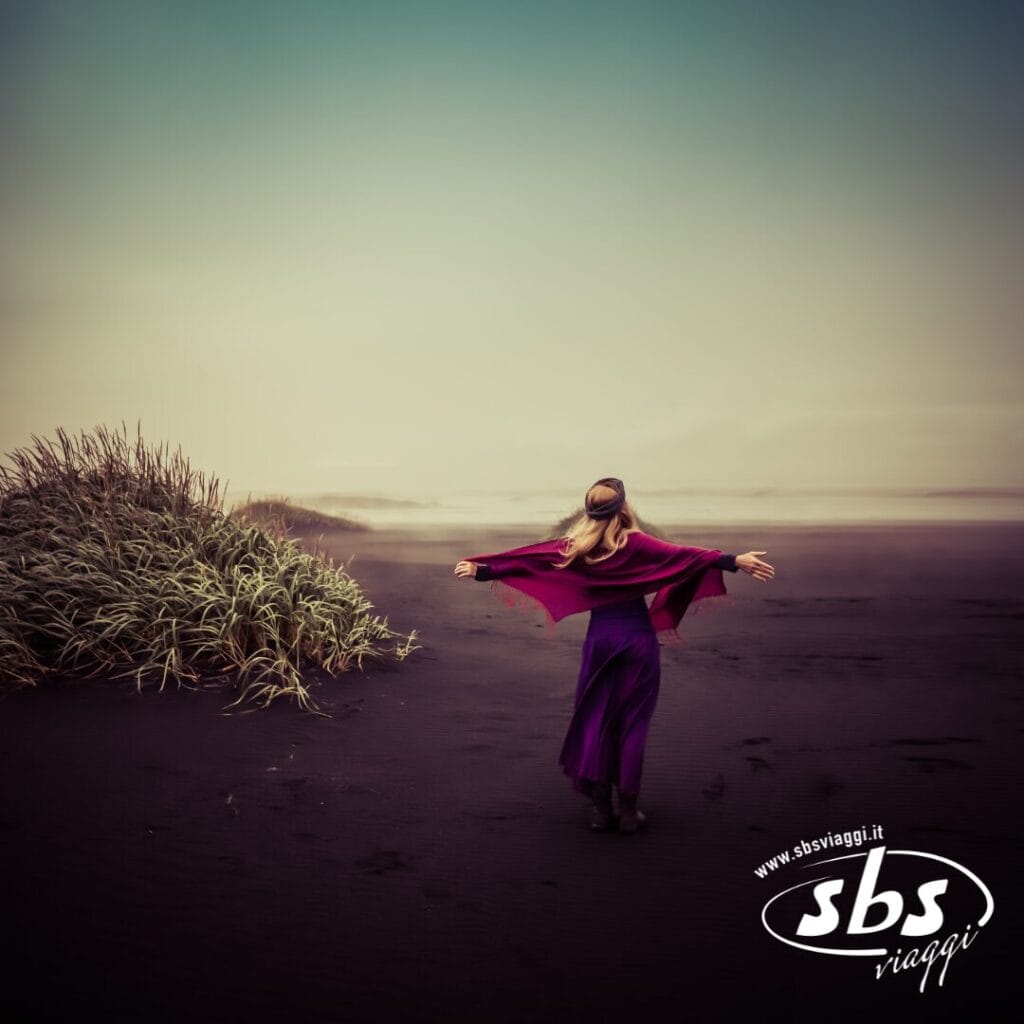 Una persona con un abito viola e uno scialle rosso è in piedi con le braccia tese su una spiaggia di sabbia nera a Sud Islanda, vicino all'erba alta; è visibile il logo di SBS Viaggi, che evoca la bellezza selvaggia delle Isole Vestman.