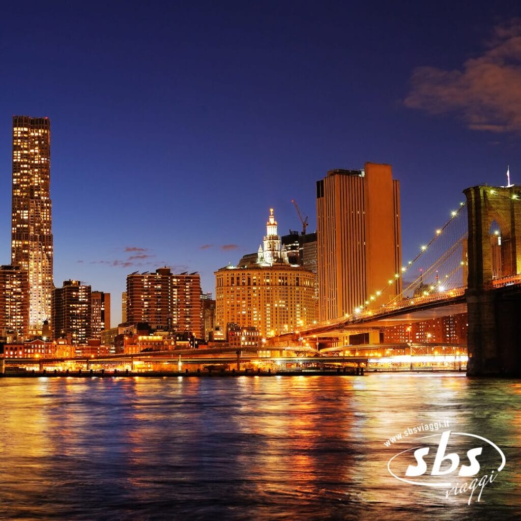 Uno scatto urbano notturno con edifici illuminati e il ponte di Brooklyn riflesso nell'acqua sotto un cielo blu scuro, che cattura la magia di Settembre a New York.
