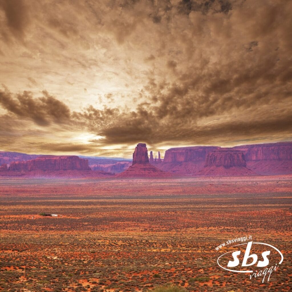 Paesaggio desertico con formazioni rocciose rosse e altopiani sotto un cielo nuvoloso e suggestivo al tramonto; logo del Tour del Golden West nell'angolo in basso a destra.