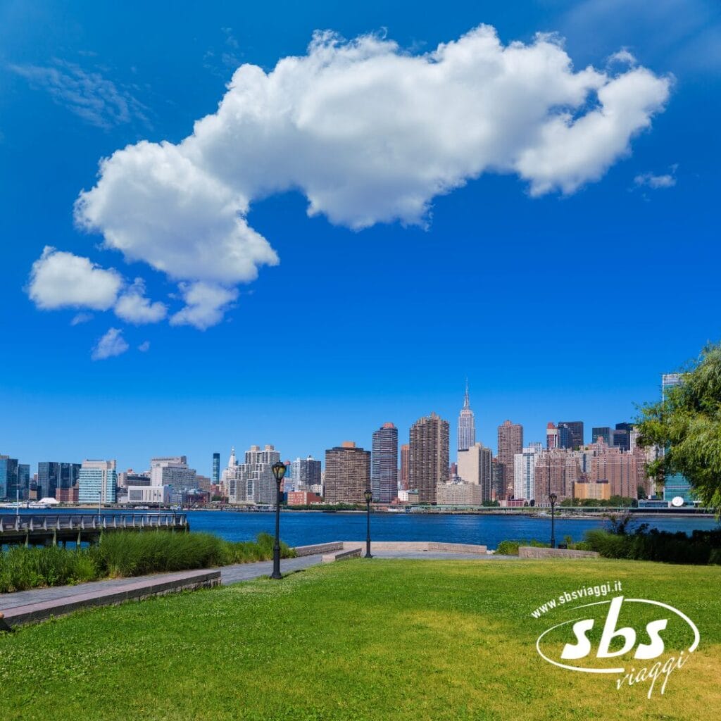 Un parco con erba verde si affaccia su un fiume e sullo skyline di New York City sotto un cielo azzurro con nuvole sparse; nell'angolo compaiono il logo