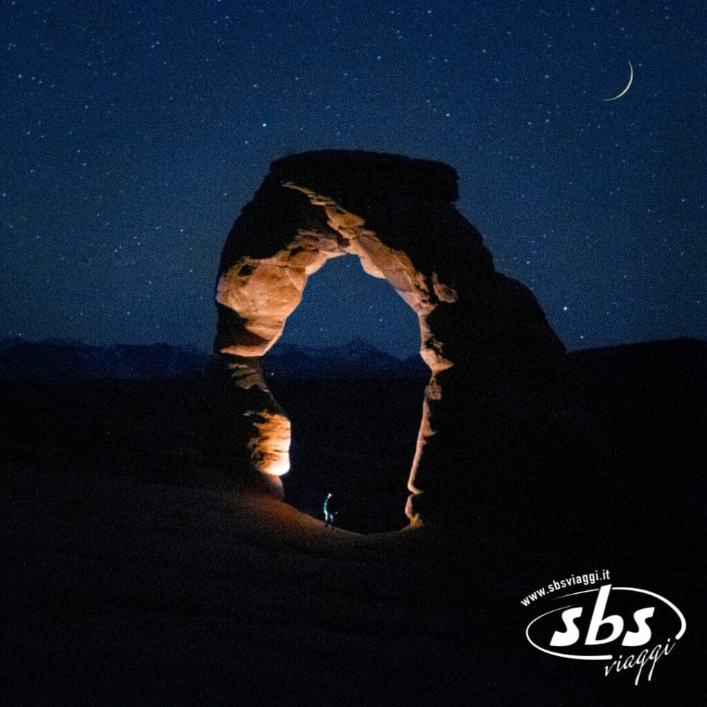 Una persona è in piedi sotto Delicate Arch di notte, illuminata da una torcia, con un cielo stellato sullo sfondo. La scena cattura sia la tradizione che la modernità, con il logo "sbs viaggi" in basso a destra.