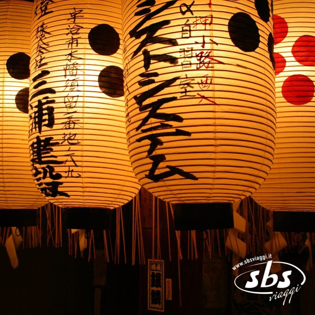 Tre lanterne di carta giapponesi illuminate con calligrafia nera e rossa emanano una calda luce, evocando lo spirito del Giappone. Il logo di SBS Viaggi è visibile nell'angolo in basso a destra, a suggerire un indimenticabile viaggio Foliage 2025.