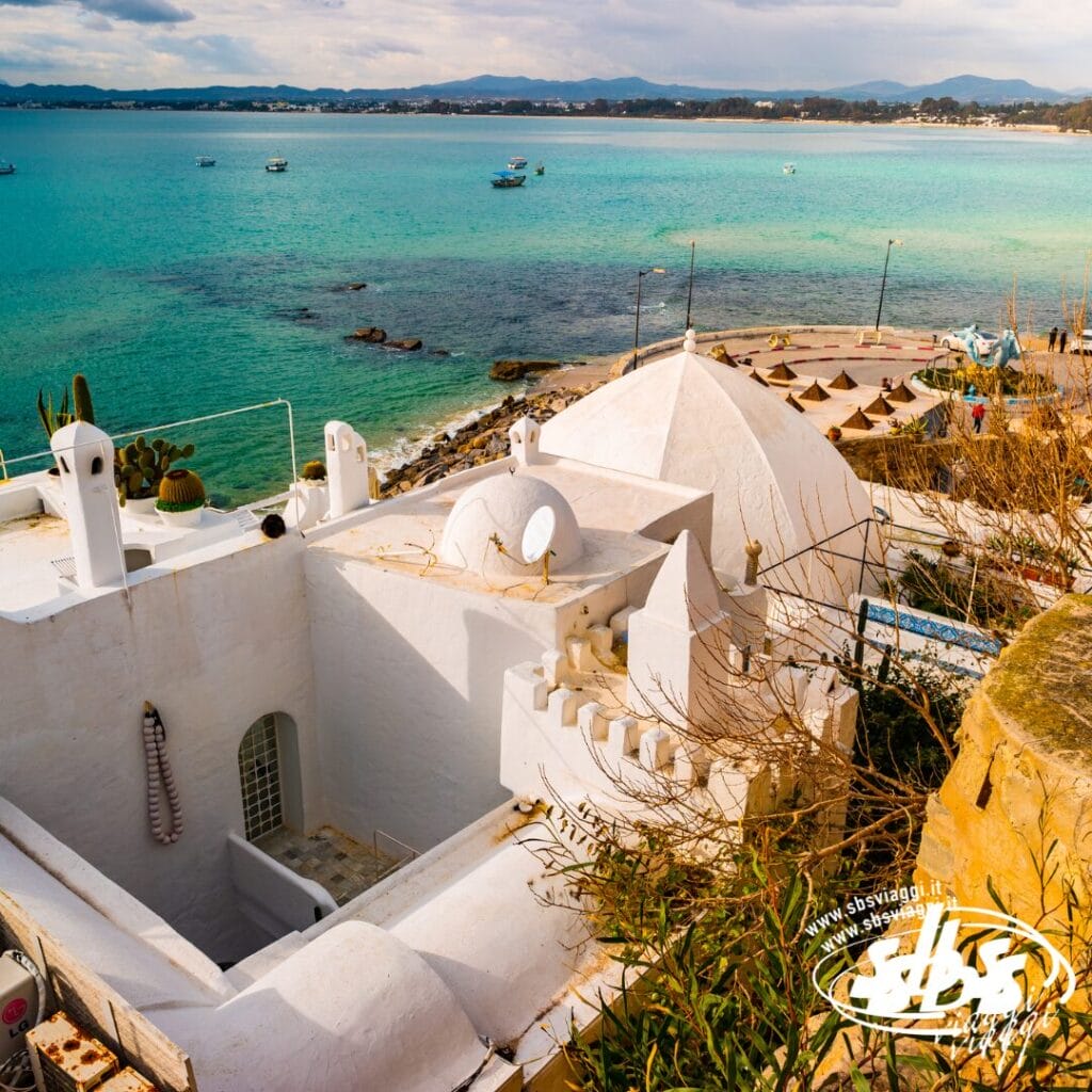 Un edificio bianco in stile mediterraneo che si affaccia sul mare turchese e sulla spiaggia, con barche e ombrelloni sotto un cielo nuvoloso: ammirate questa vista durante il Tour Autentica Tunisia.