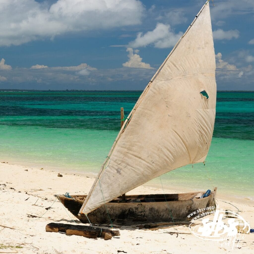 Un piccolo dhow di legno con una vela bianca è adagiato su una spiaggia sabbiosa nei pressi delle acque turchesi del Mare, catturando la tranquilla bellezza della Tanzania e Mare.