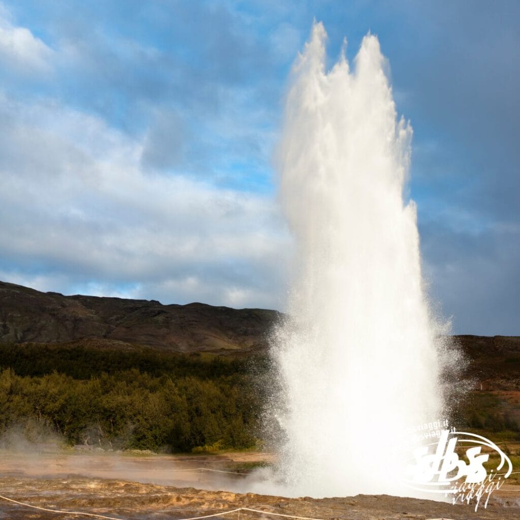 Un grande geyser erutta in Islanda, sparando acqua in aria contro un cielo nuvoloso e un paesaggio verde: uno spettacolo maestoso durante un Viaggio Astronomico.