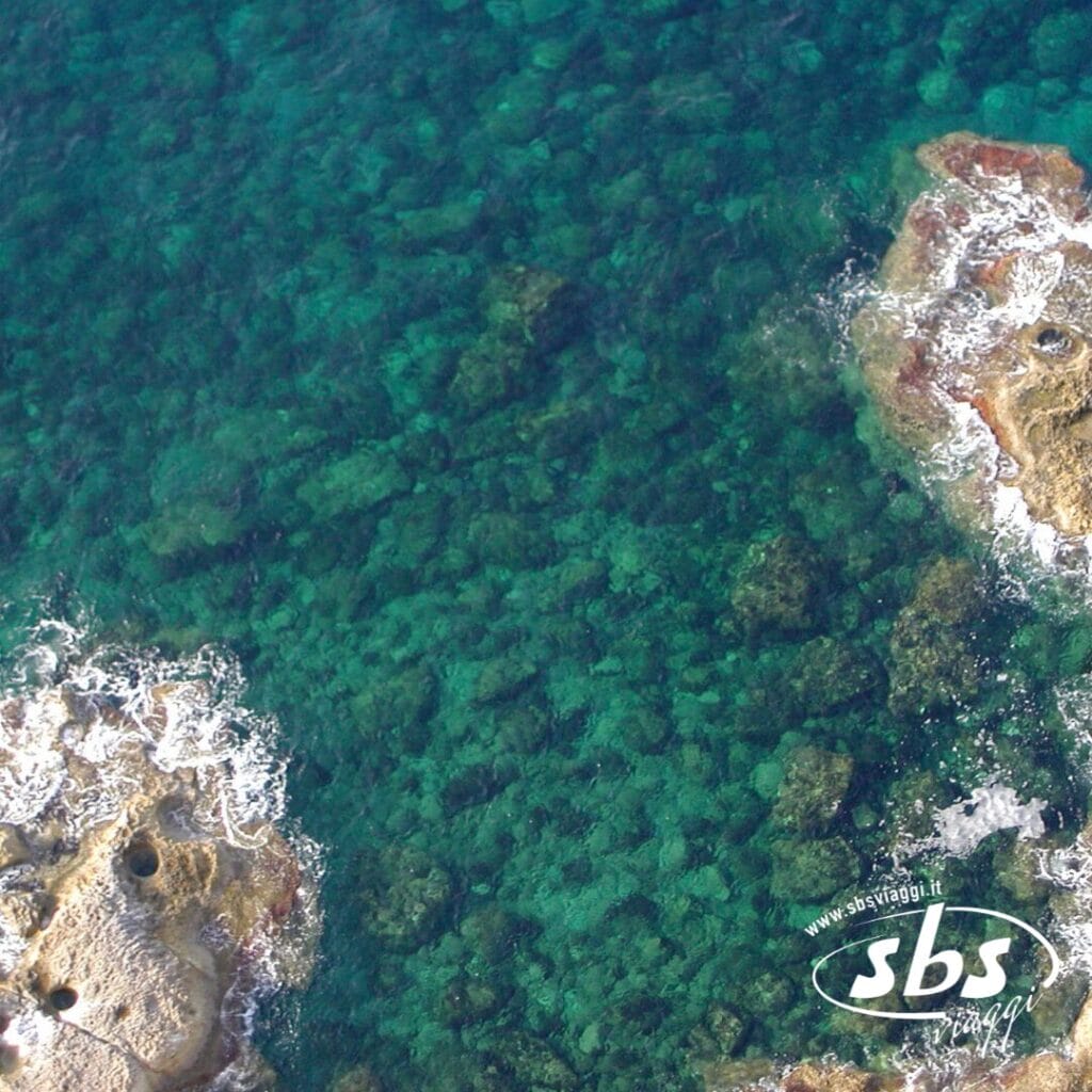 Vista aerea del mare cristallino color turchese con formazioni rocciose e schiuma bianca in superficie, a pochi passi da Demo Villaggi e Demo Hotel.