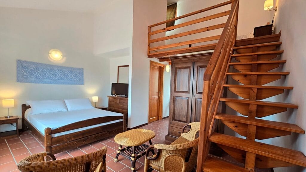 Un'accogliente camera d'albergo con soppalco in legno, letto matrimoniale, sedie in vimini e scala in legno offre la perfetta combinazione di relax e comfort.