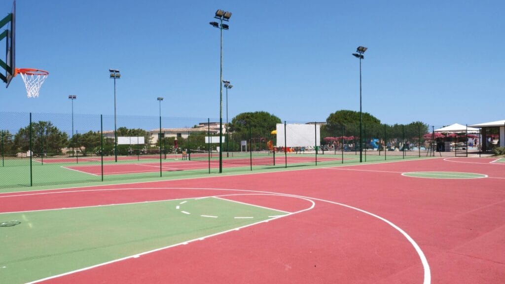 Campi da basket e da tennis all'aperto con superfici rosse e verdi, circondati da recinti e alberi, sotto un cielo limpido: un luogo perfetto per gli amanti dello sport che vogliono godersi una partita o provare una nuova Bozza automatica.
