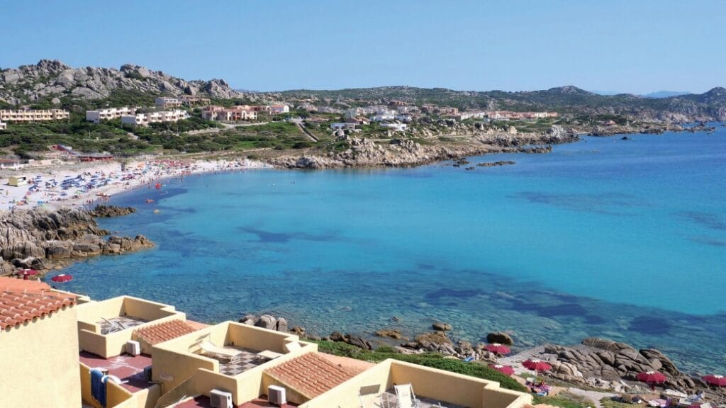 Una città costiera con acque cristalline, una costa rocciosa, una spiaggia sabbiosa e edifici in collina sotto un cielo terso creano una Bozza automatica da cartolina.