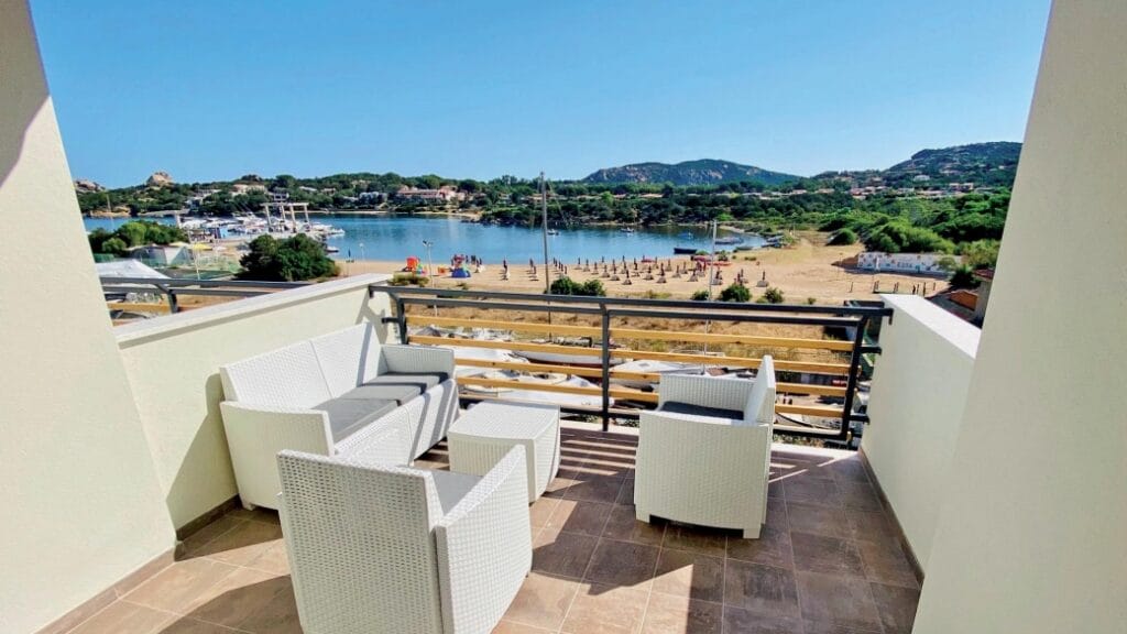 Un balcone con sedie bianche si affaccia su una spiaggia sabbiosa, acqua cristallina e verdi colline nelle giornate di sole: perfetto per rilassarsi o godersi la brezza della Bozza automatica.