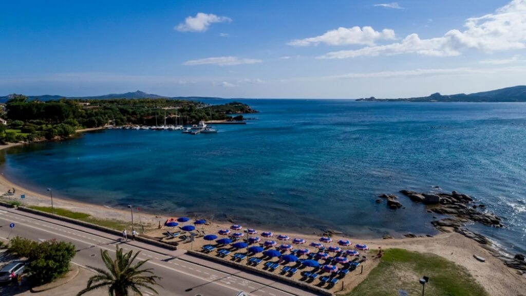 Una spiaggia soleggiata con ombrelloni blu, acqua limpida, una strada e in lontananza verdi colline sotto un cielo azzurro offrono la cornice perfetta per una rilassante fuga in compagnia di una bozza automatica.